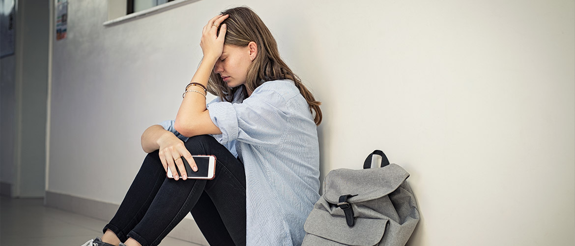 Jeune fille en détresse assise contre un mur, tenant un smartphone, symbolisant les conséquences émotionnelles du cyberharcèlement ou de la sextorsion chez les adolescents